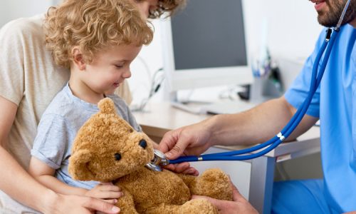 Portrait of adorable curly child  sitting on mothers lap in doctors office holding teddy bear toy, with pediatrician listening to heartbeat using stethoscope